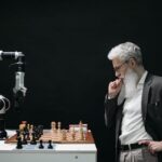 An elderly scientist contemplates a chess move against a robotic arm on a chessboard.
